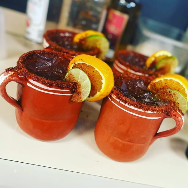 Four Mexican micheladas in red clay mugs, garnished with chili powder, orange, and lime slices.