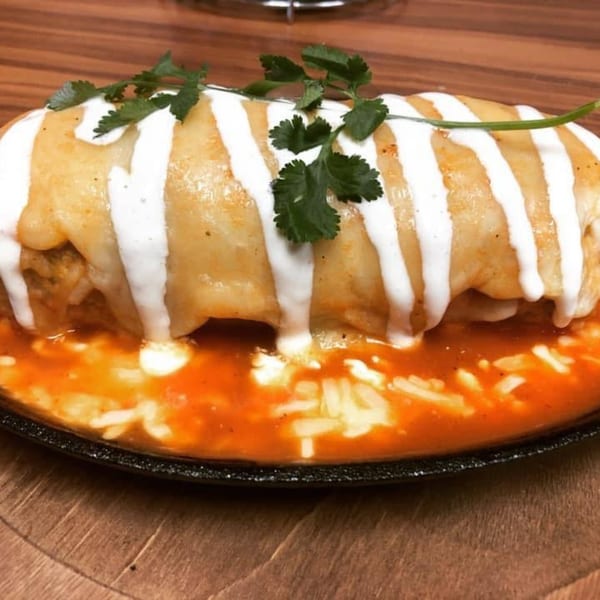 A large, golden-brown burrito covered in white crema and cilantro, sitting in a red sauce.