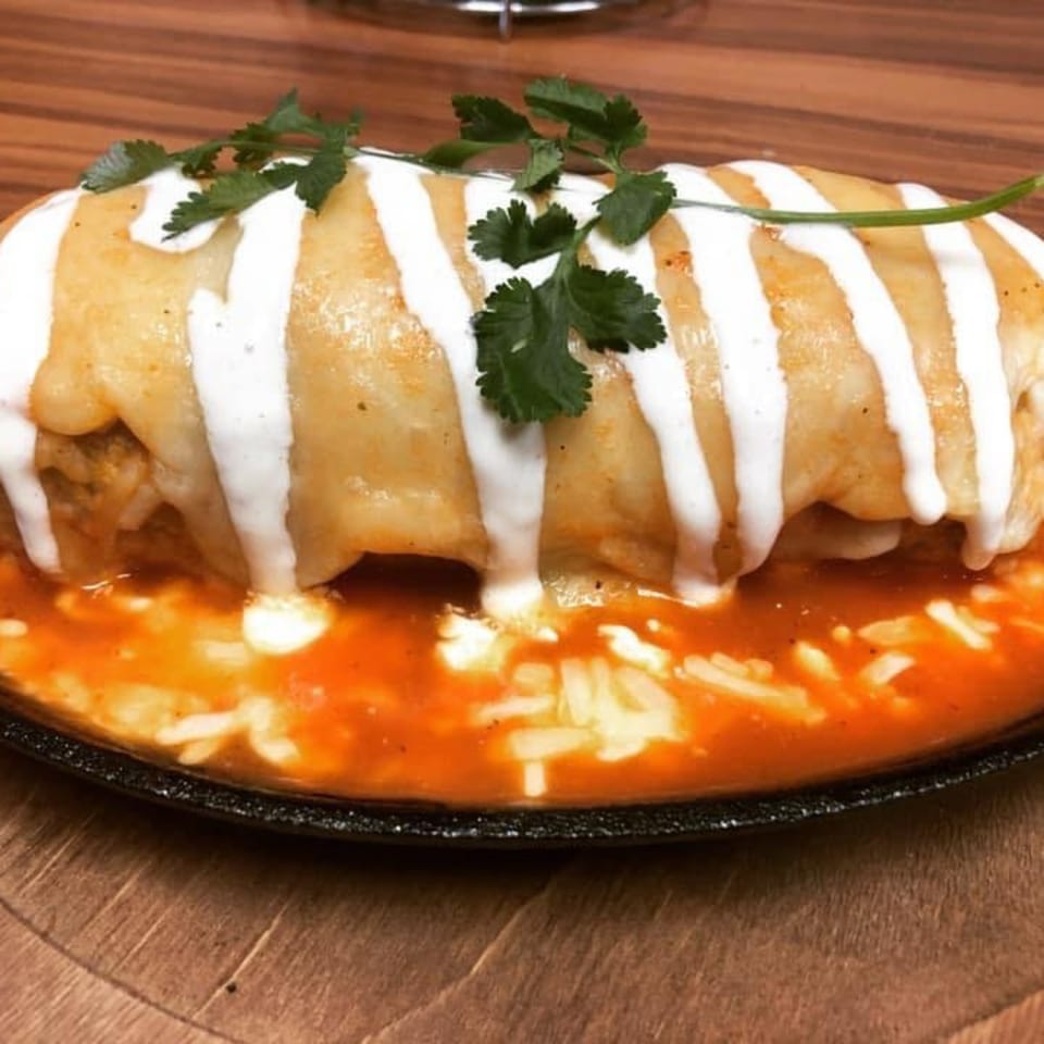 A large, golden-brown burrito covered in white crema and cilantro, sitting in a red sauce.