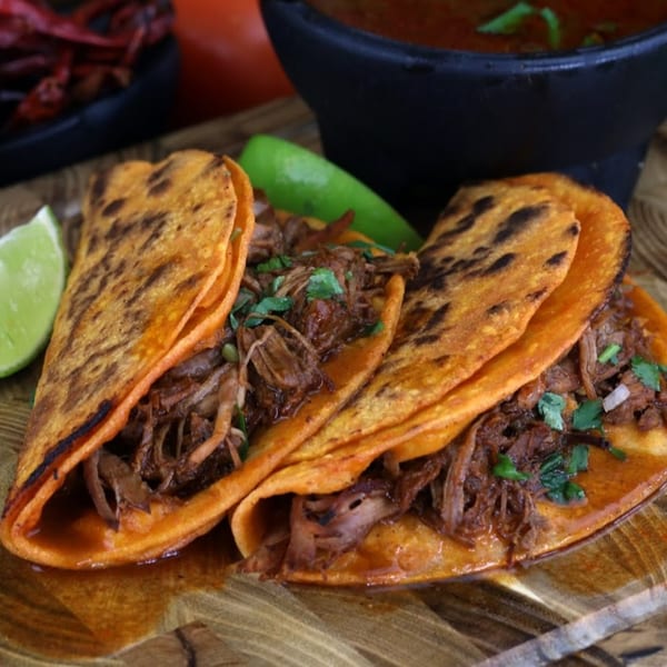 Three Pika Tortas Ahogadas birria tacos with lime wedges and a bowl of consommé.